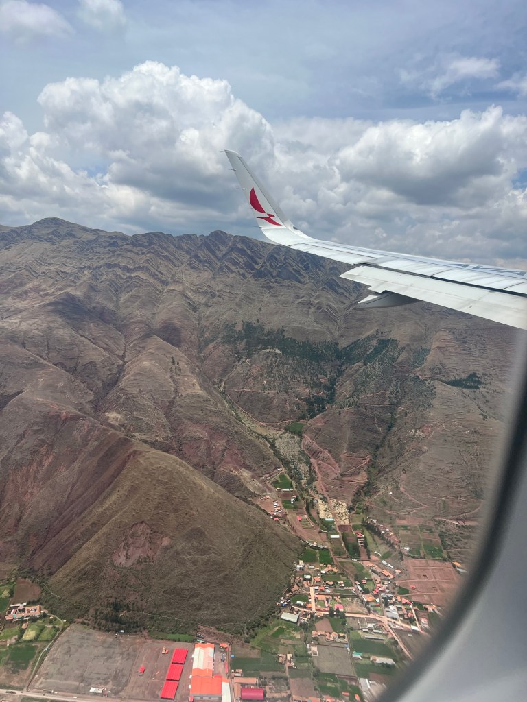 View from an airplane window showing rugged mountains and valleys below with a plane wing in the corner, indicating descent into Cusco, Peru.