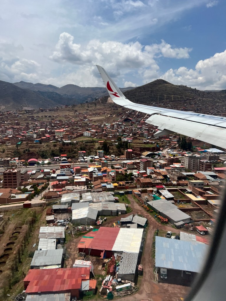 An airplane wing with a logo flying above a colorful landscape of houses and hills in Cusco, Peru.