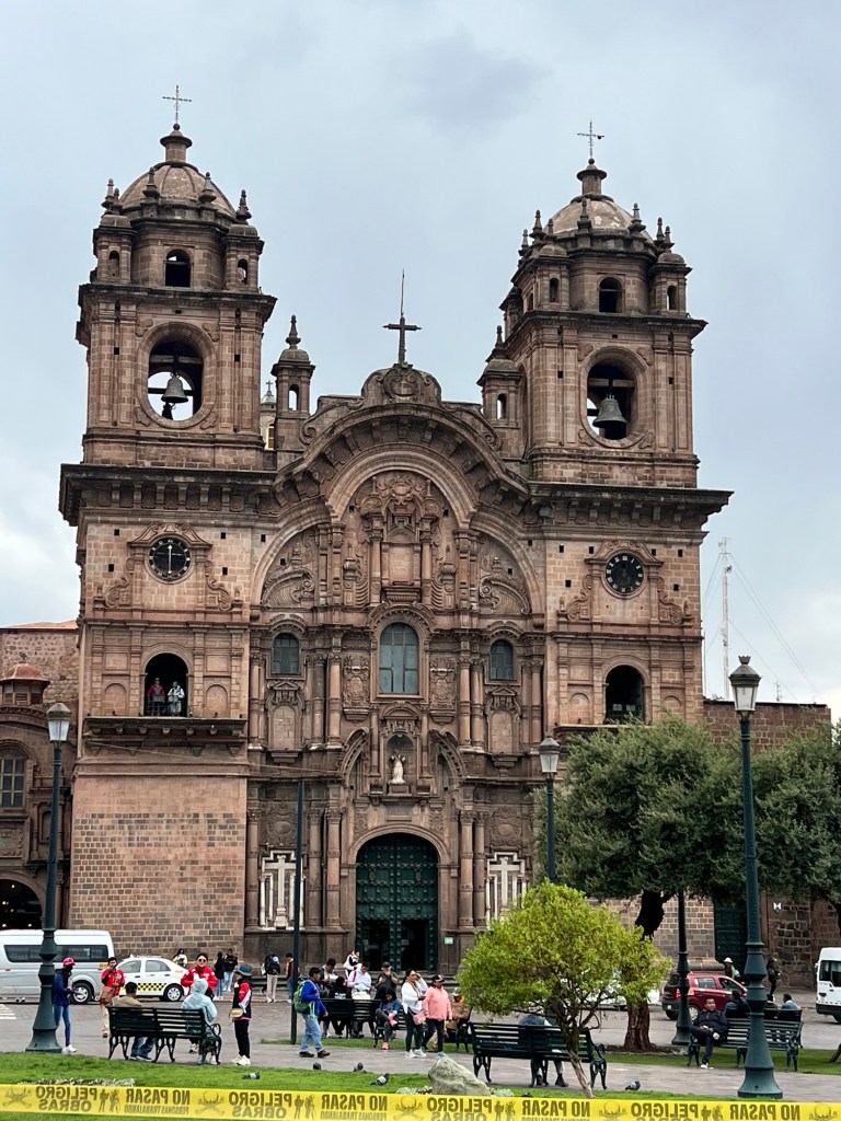 A historic church building in Cusco, Peru, featuring ornate architectural details, bell towers, and a green entrance, surrounded by people and benches in a plaza.
