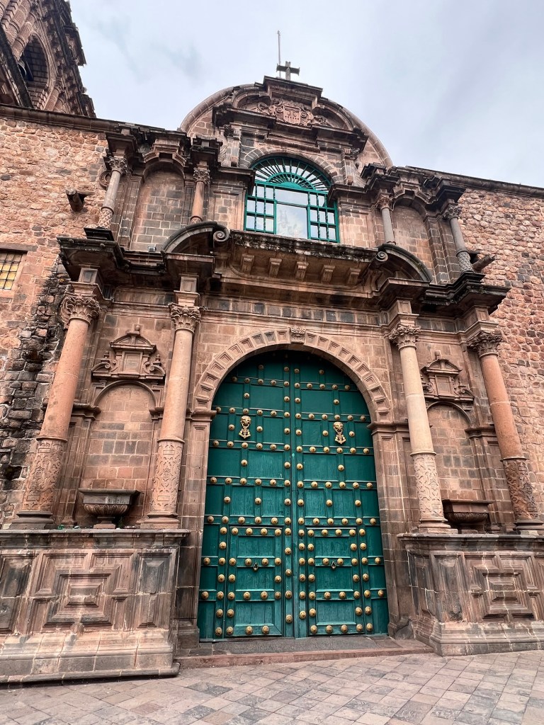 A close-up view of a historic building entrance in Cusco, Peru, featuring a large green wooden door adorned with decorative brass knobs, framed by intricate stone architecture.
