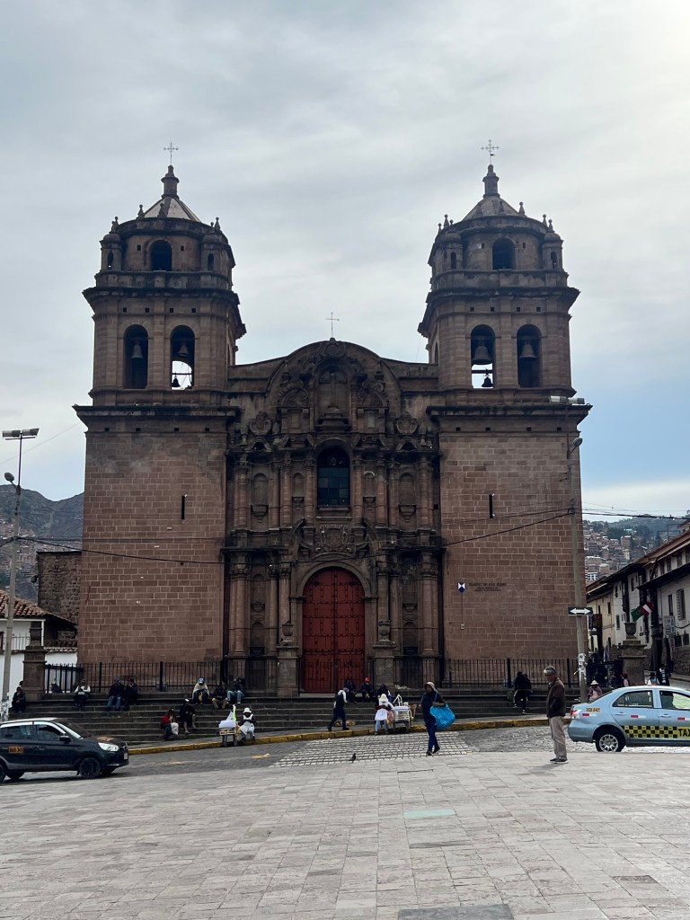 Front view of a historic church in Cusco, Peru, featuring two bell towers and a decorative facade, with people and cars around.