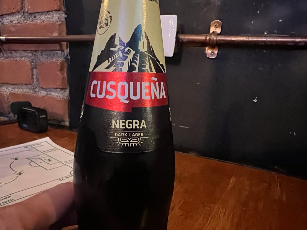 A close-up image of a bottle of Cusqueña Negra dark lager beer on a wooden table, with a brick wall in the background.