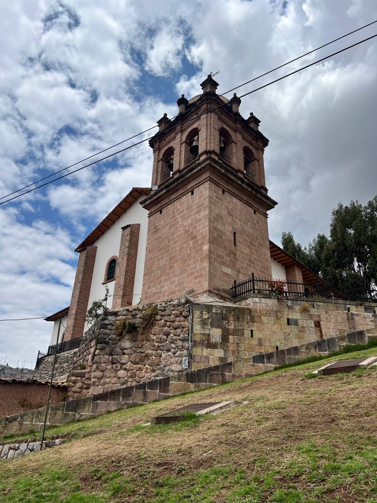 A stone church with a bell tower located on a hillside, surrounded by green grass and trees under a partly cloudy sky.