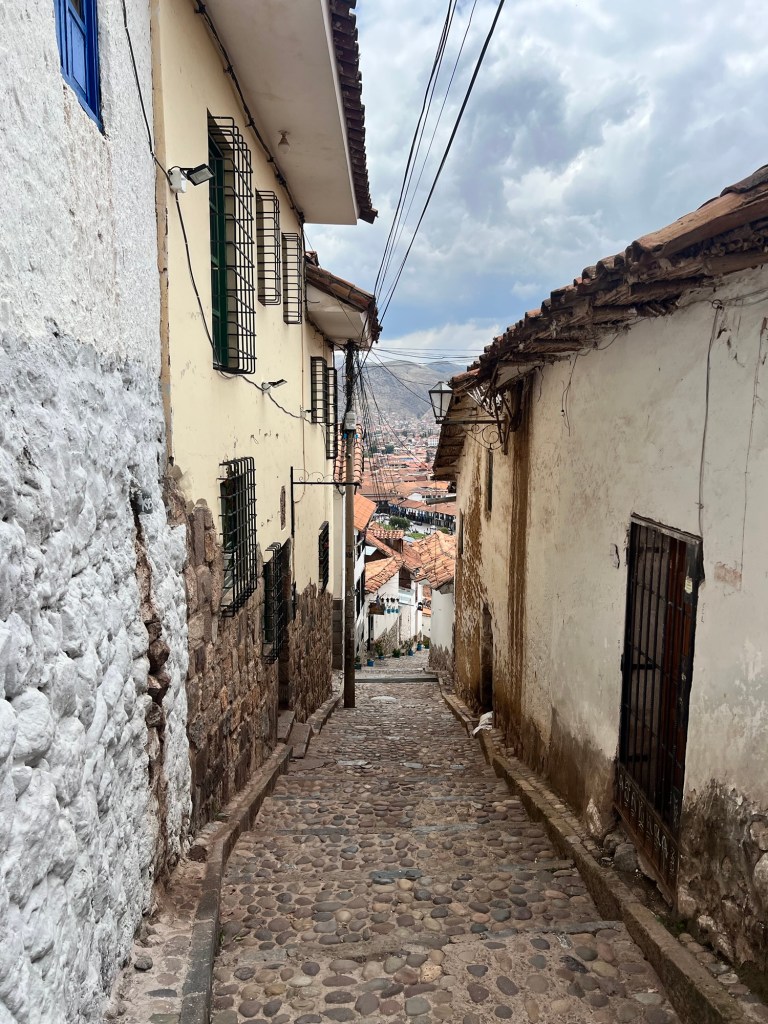 Narrow cobblestone street in Cusco, Peru, lined with whitewashed buildings and colorful rooftops in the distance.