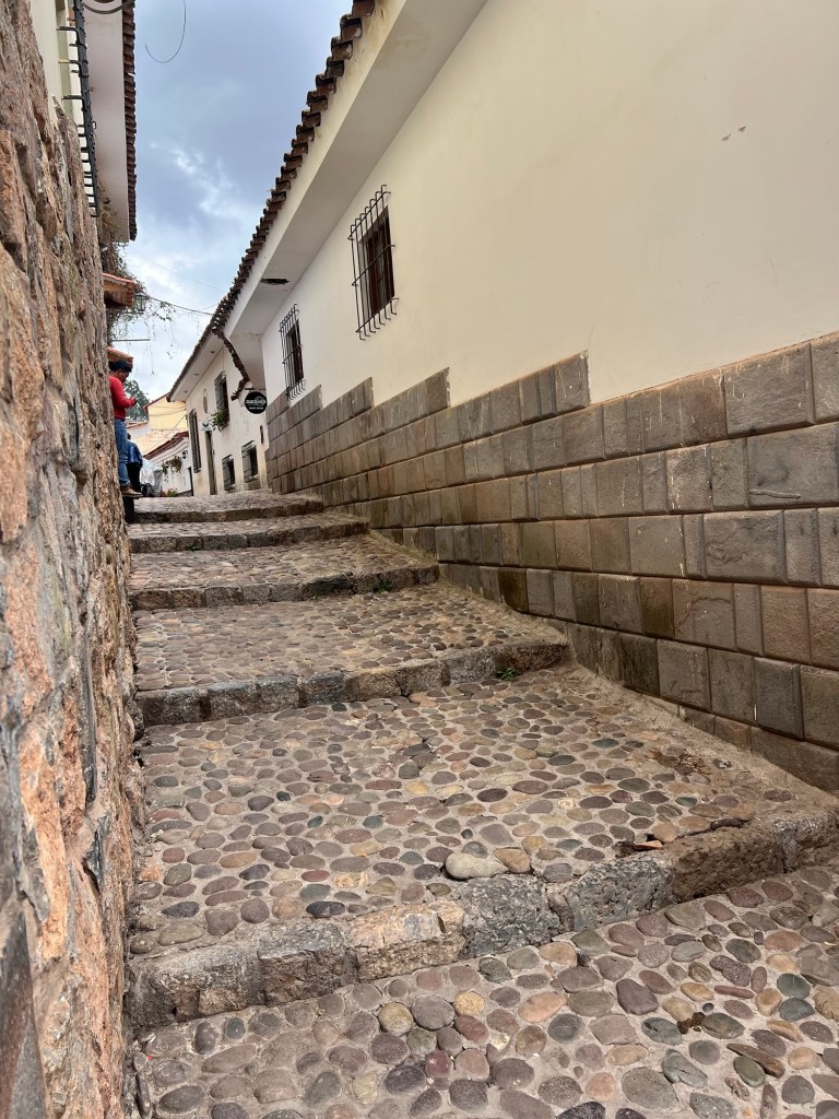 A narrow cobblestone street with steps leading up, flanked by whitewashed buildings and dark stone walls, under a cloudy sky.