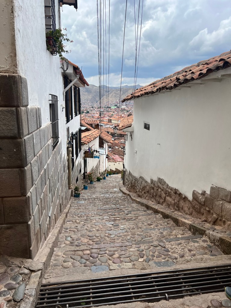 A narrow cobblestone street in Cusco, Peru, lined with white-walled houses and flower pots, leading downhill with a view of the city and mountains in the distance.