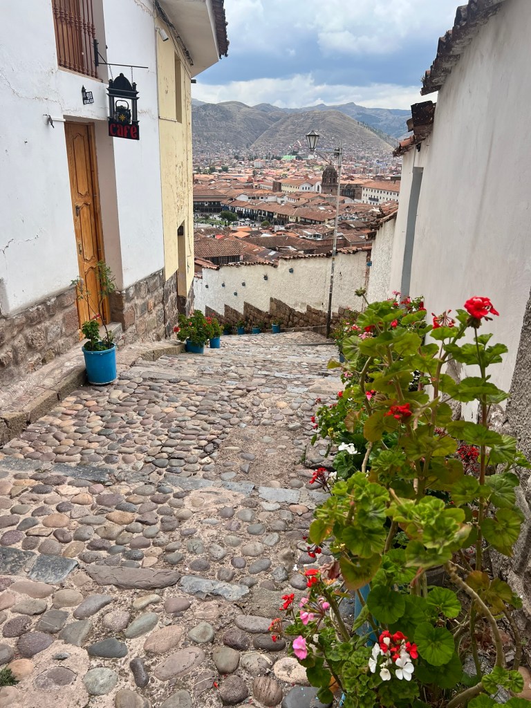 A cobblestone street in Cusco, Peru, with colorful flower pots lining the sides and a view of the city below. The scene captures traditional architecture and the surrounding mountains.