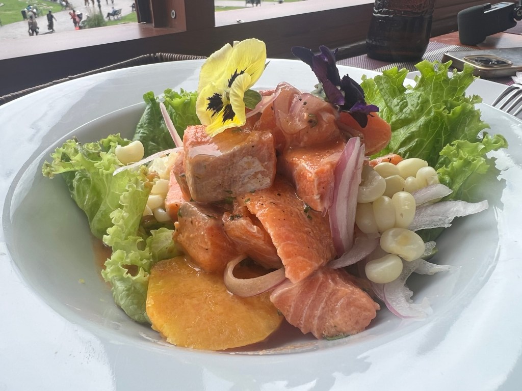 A vibrant plate of ceviche featuring salmon chunks, garnished with fresh lettuce, onions, and edible flowers, surrounded by a mix of vegetables and accompanied by a side of traditional corn.