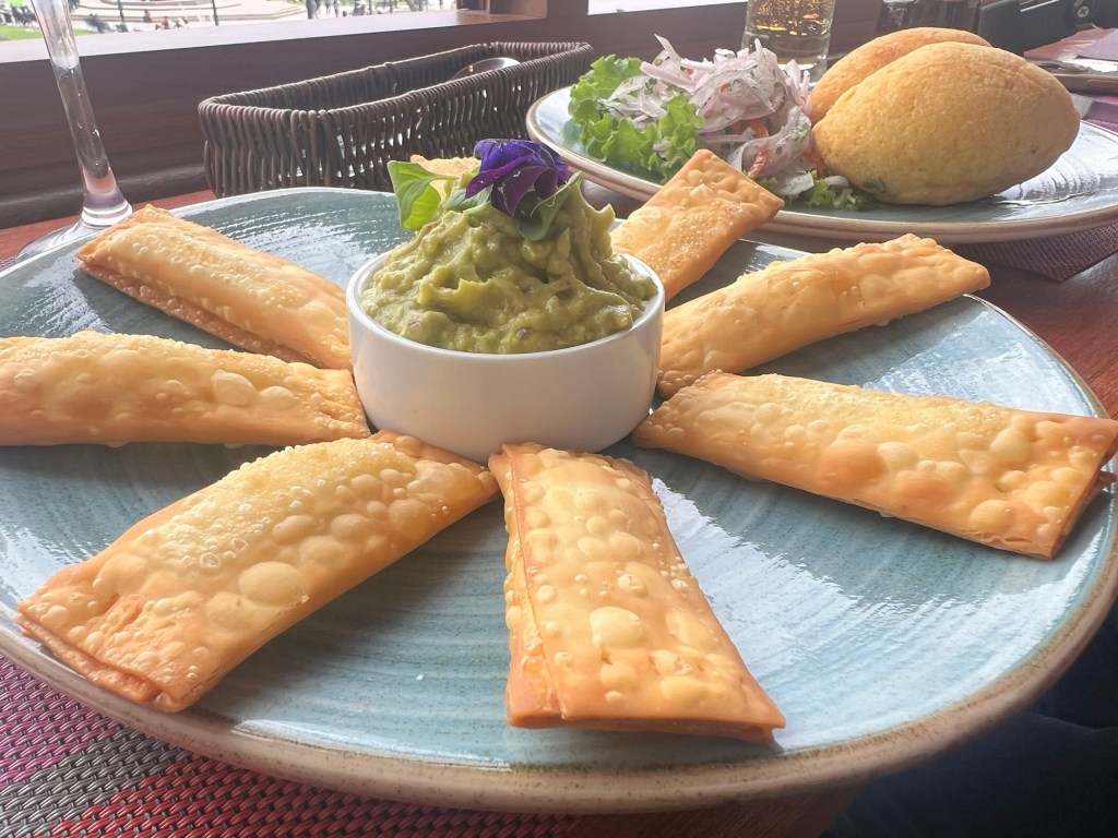 A plate of golden empanadas arranged in a circular pattern around a small bowl of guacamole, accompanied by a side salad and two bread rolls.