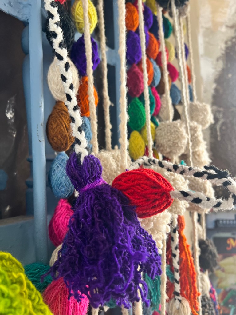Close-up of colorful handmade yarn decorations, featuring various vibrant pom-poms and tassels in shades of purple, red, blue, yellow, and green, displayed against a blue background.