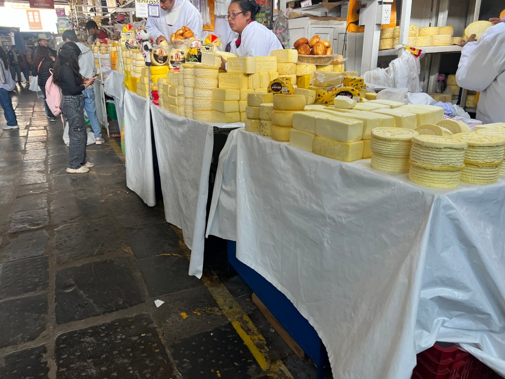 A cheese stall at a market in Peru, featuring various types of stacked cheeses and vendors in white aprons attending to customers.