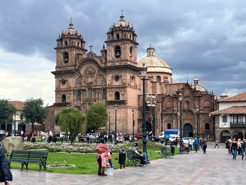 A historic church in Cusco, Peru, featuring intricate architecture and twin towers, surrounded by people and greenery in a lively square.