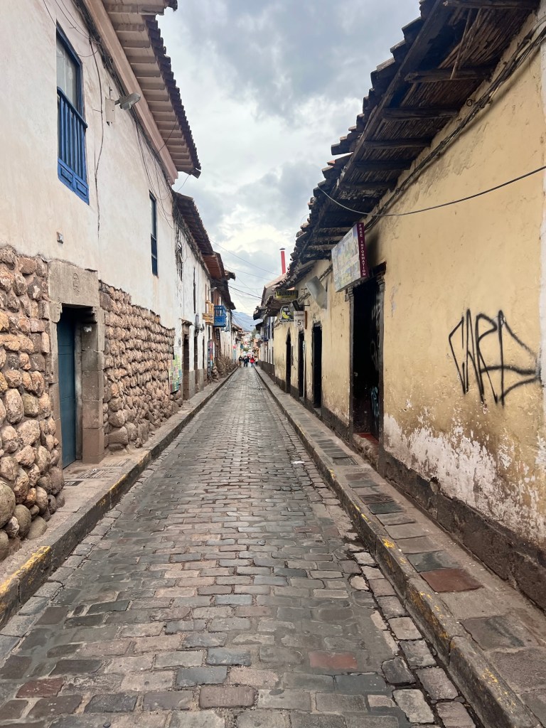 A cobblestone street in Cusco, Peru, lined with rustic buildings featuring traditional architecture and colorful doorways under a cloudy sky.