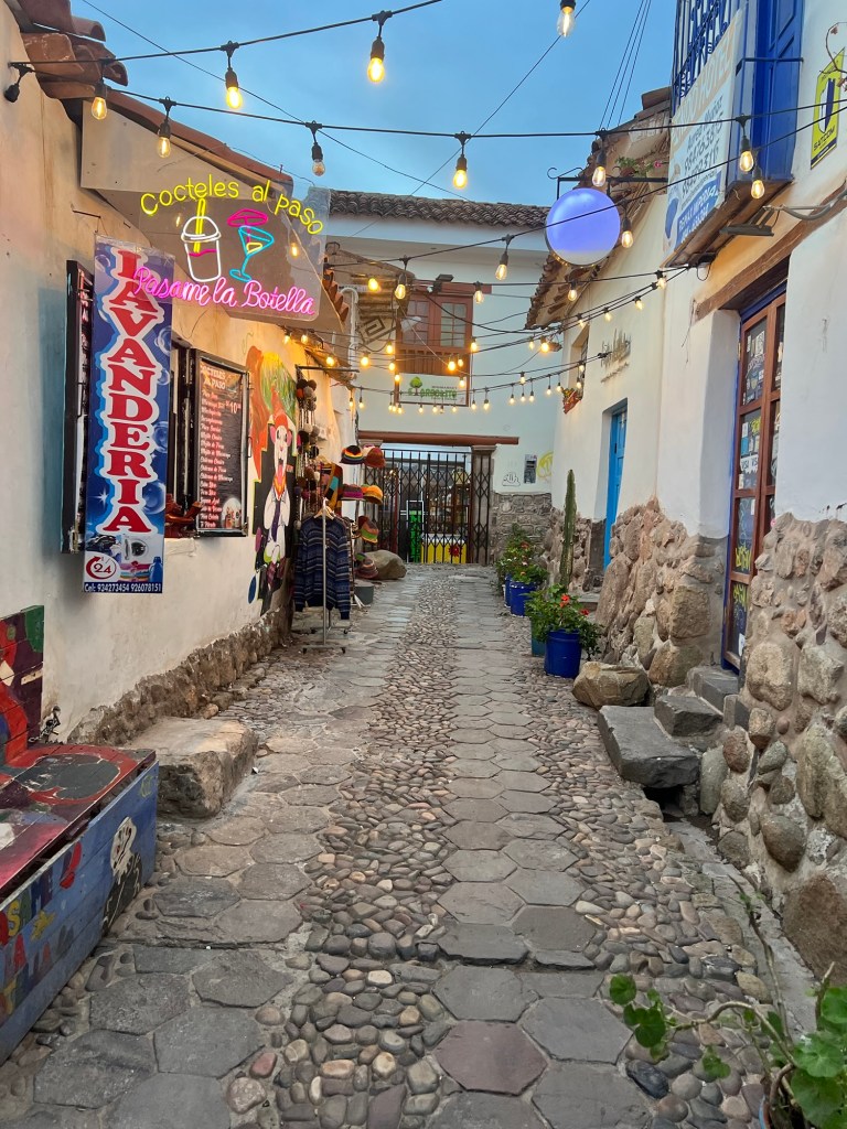 A narrow street in Cusco, Peru, lined with colorful buildings and string lights, featuring signage for local businesses.
